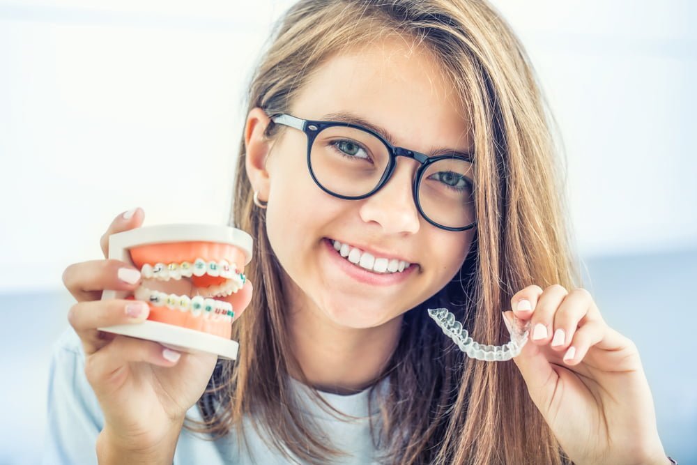 Smiling teenage girl with glasses holding a dental model with braces in one hand and a clear Invisalign aligner in the other, representing orthodontic options – Invisalign Beverly Hills