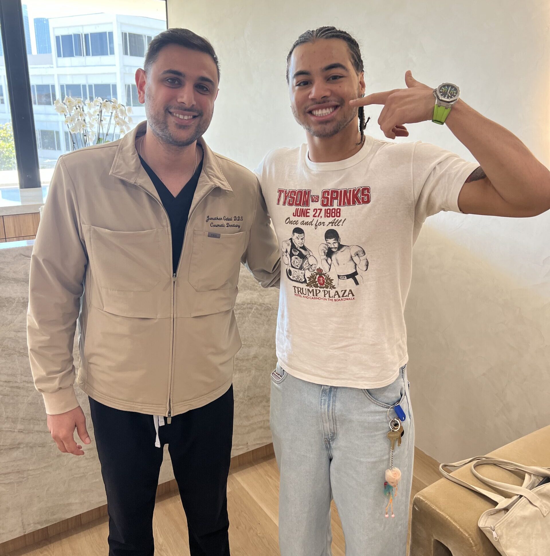 Young man smiling and pointing to his teeth while standing next to his dentist in a modern dental office, celebrating Invisalign success – Invisalign Beverly Hills 