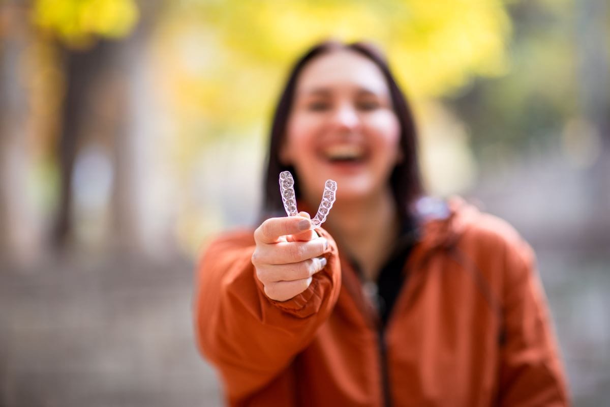 Smiling woman outdoors holding up Invisalign clear aligners toward the camera, showcasing orthodontic treatment in a casual setting.