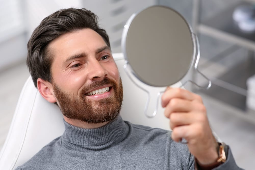 Bearded man smiling while looking into mirror after dental makeover – Porcelain Veneers Los Angeles
