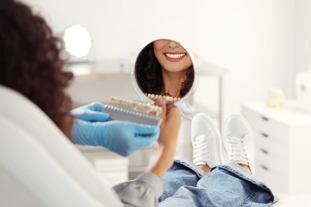 Smile Evaluation with Shade Guide – Porcelain Veneers Beverly Hills Patient admiring her smile in a handheld mirror while a dental professional matches veneer shades using a color guide – Porcelain Veneers Beverly Hills