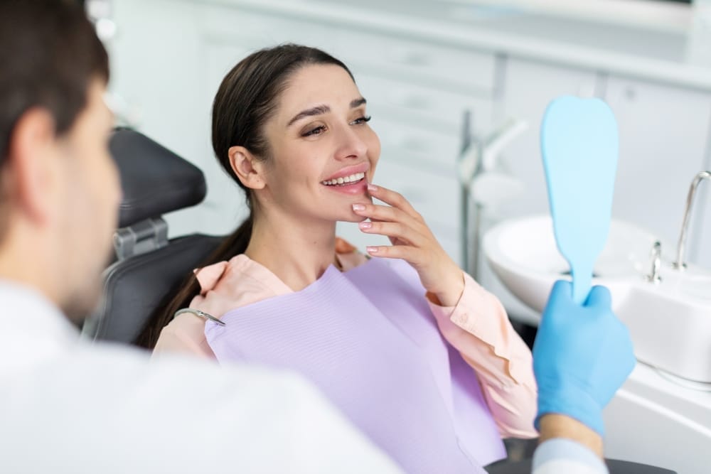 Young woman smiling at her reflection in a handheld mirror during a dental visit, showcasing results of teeth whitening in Beverly Hills