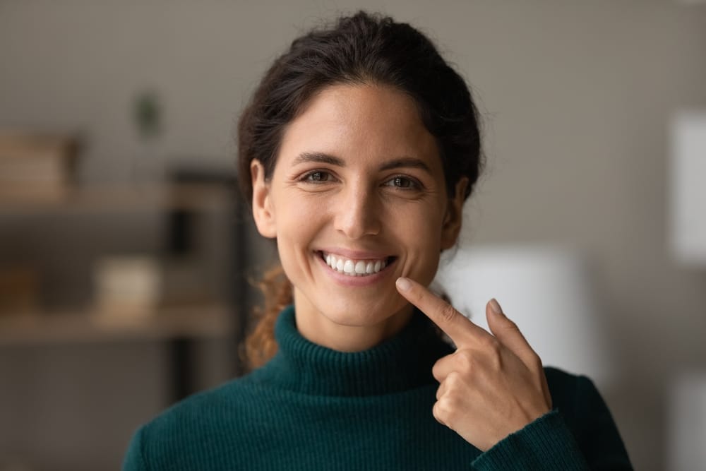 Natural Smile Results After Veneers  Brunette woman in green sweater smiling and pointing to her bright, natural-looking teeth, showcasing successful porcelain veneers
